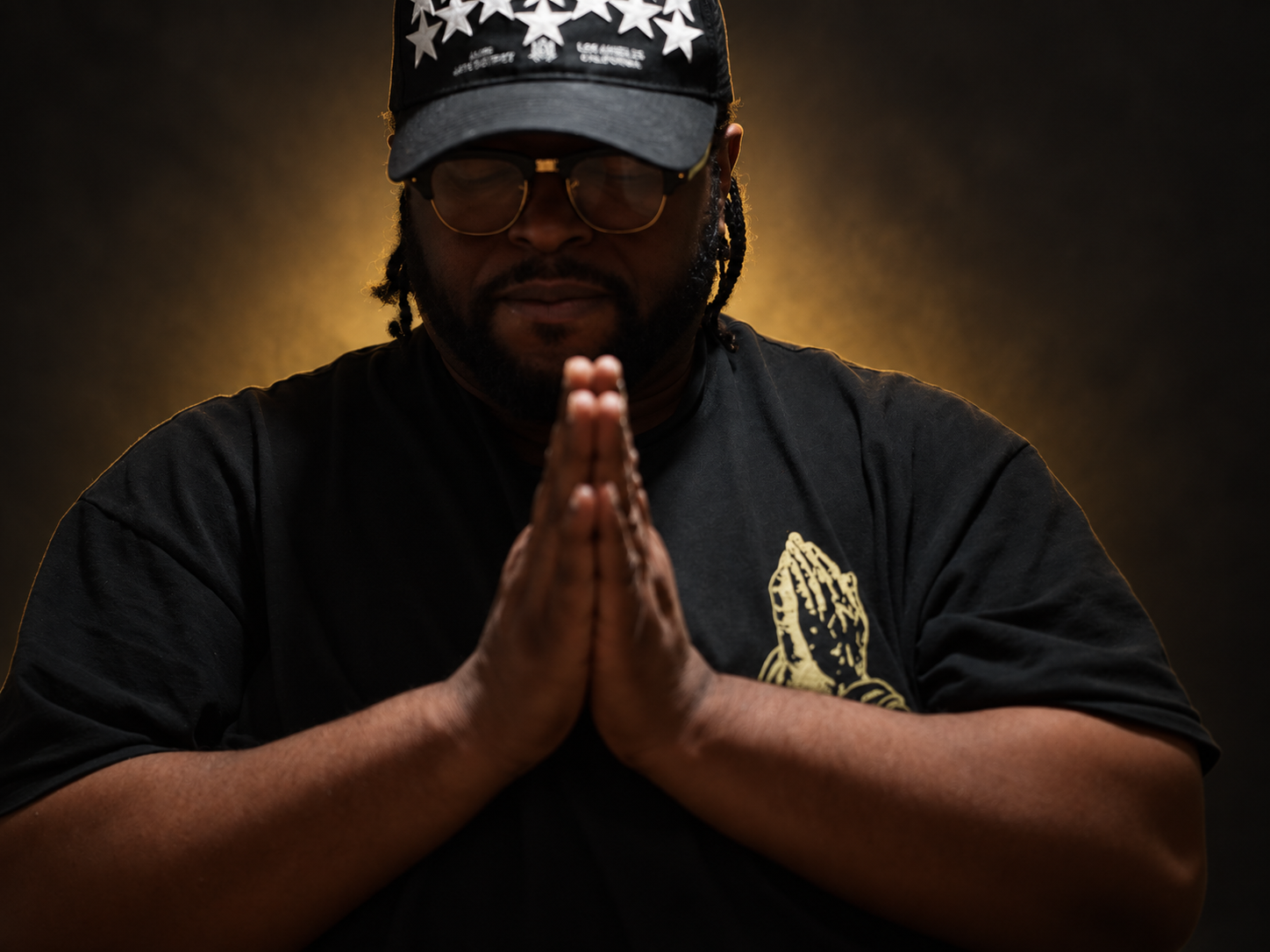 Founder of Rich Row Music wearing his signature cap and glasses, hands together, against a charcoal and gold backdrop